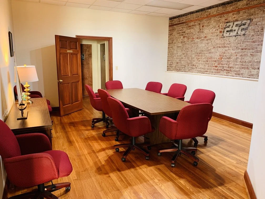 Conference room with wooden table and eight red office chairs, brick accent wall, hardwood floor, and a side table with lamp and decorative items.