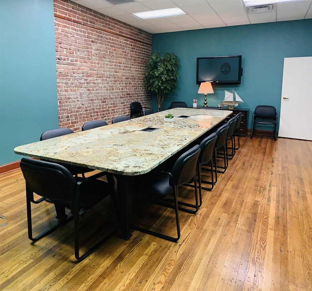 Conference room with a large granite table, black chairs, a brick wall on one side, a teal wall with a mounted flat-screen TV, a potted plant, a side table with a desk lamp, a small sailboat model, and a white door.