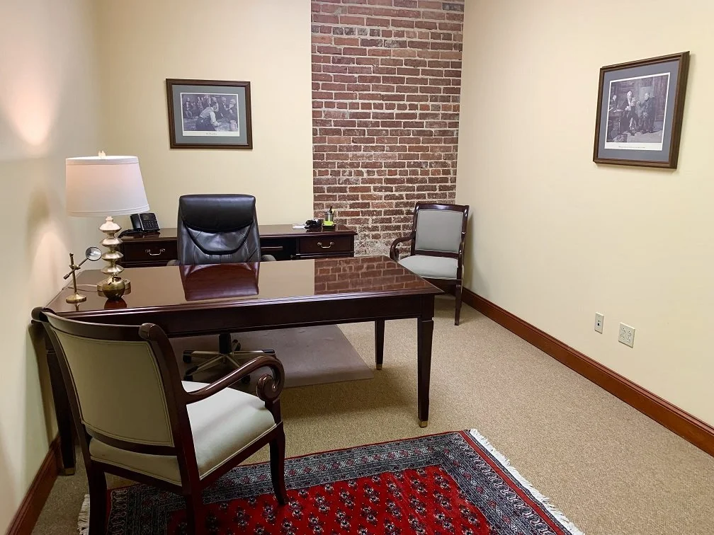 An office with a dark wood desk, a black leather office chair, two additional chairs, a lamp, a small table, a framed picture on each wall, and a red patterned rug in the foreground.