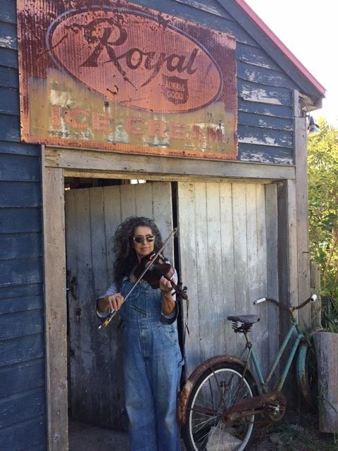 A woman with curly dark hair, sunglasses, and wearing denim overalls, plays a violin outside a rustic wooden garage or shed with a faded sign above that reads 'Royal Ice Cream'. A vintage bicycle leans against the shed nearby, with trees and foliage in the background.