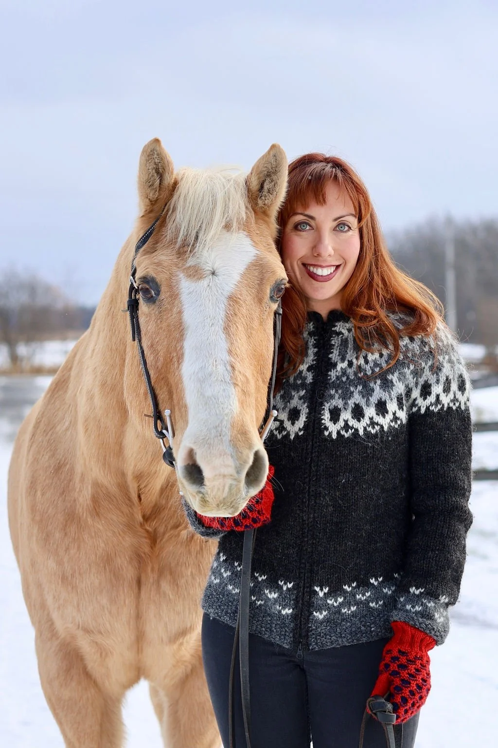 A woman with red hair smiling and holding a light brown horse with a white blaze on its face outdoors in a snowy landscape.
