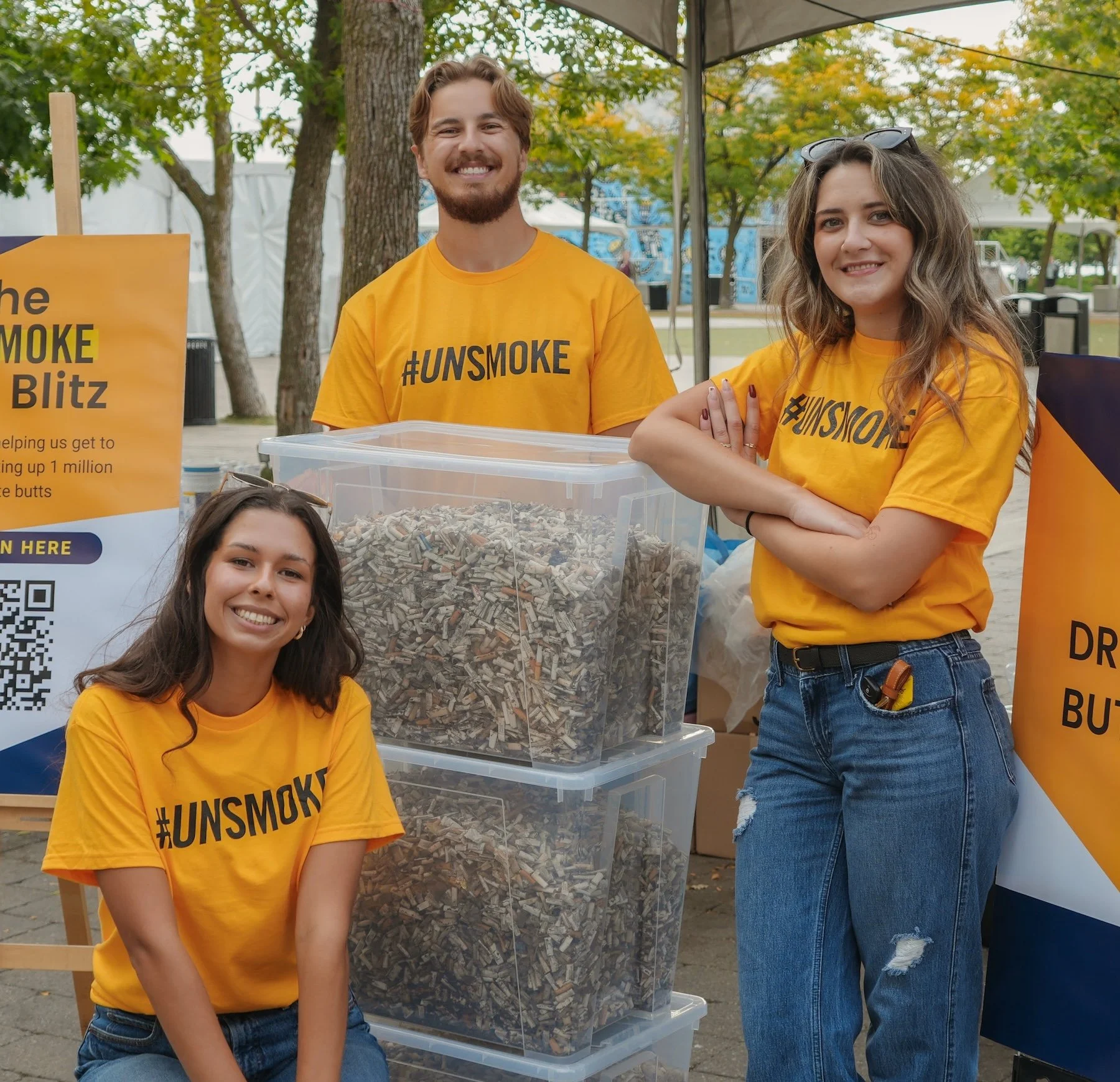 Recia, Greg and Nino at the Toronto Butt Blitz 2025 at Harbourfront Centre. They're wearing orange unsmoke t-shirts and posing with bins full of thousands of cigarette butts.