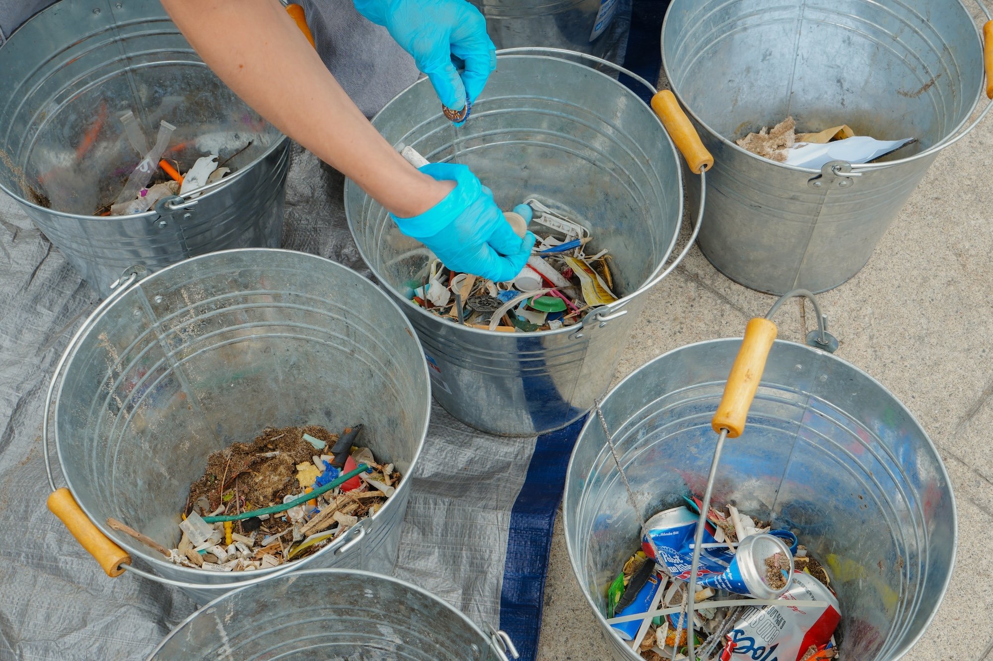 Someone wearing blue gloves sorting trash into five metal buckets filled with mixed waste, including plastic, paper, and miscellaneous debris, on a concrete surface.