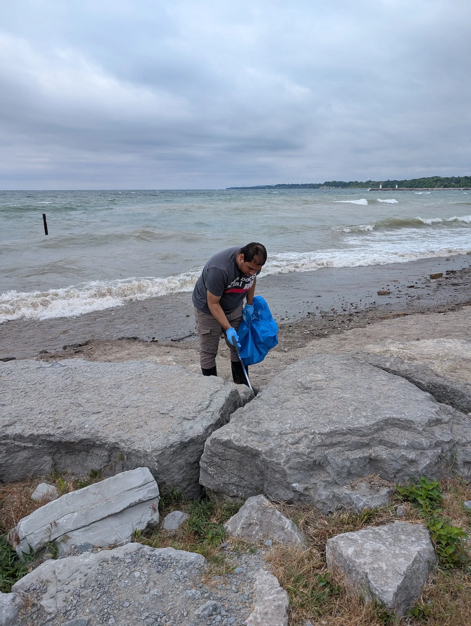 A man wearing gloves and gray clothing is collecting litter from the rocks near the shoreline of a beach on a cloudy day.