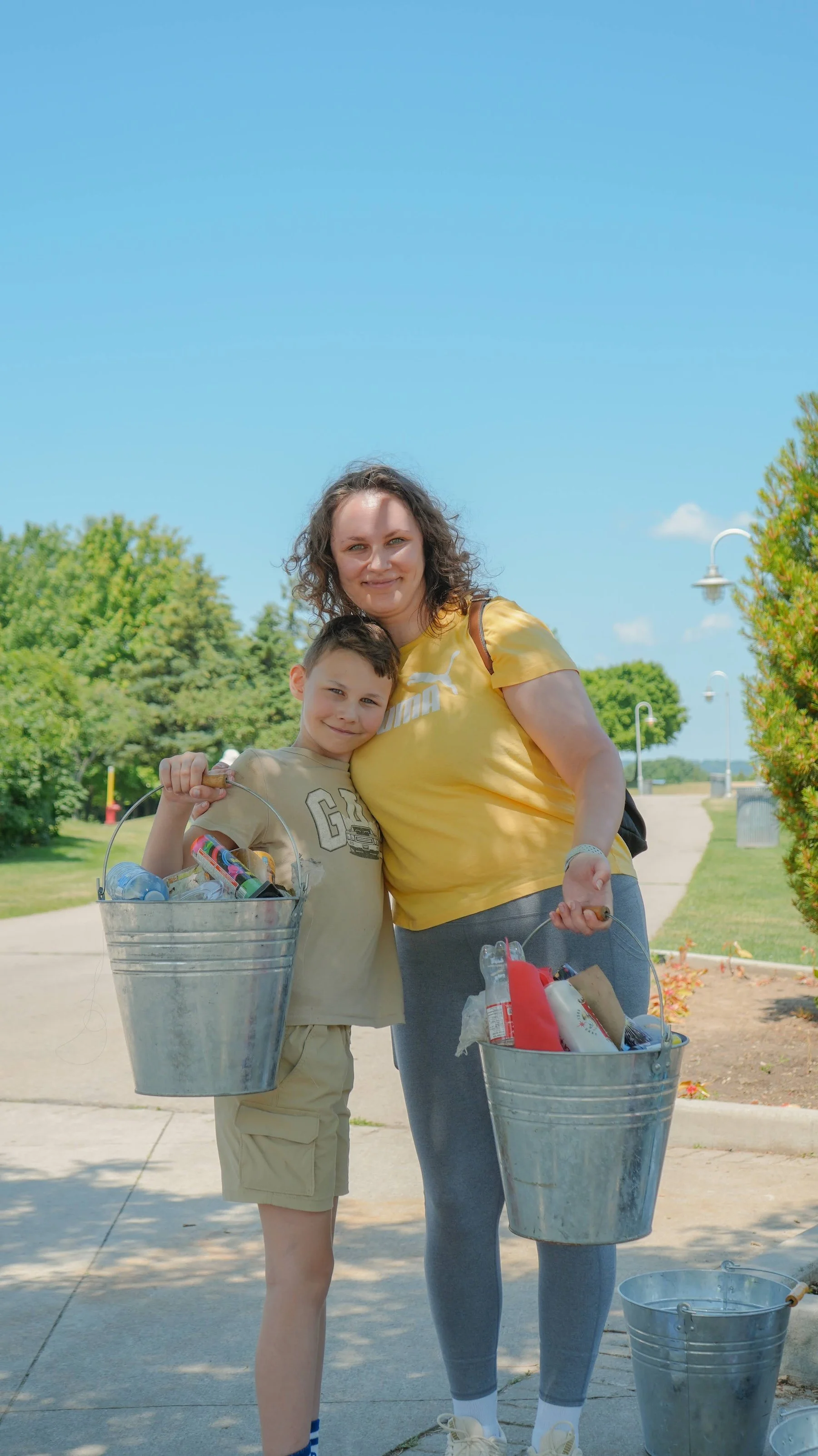 A woman and a boy holding metal buckets filled with trash, standing outdoors on a sunny day in a park or green space.