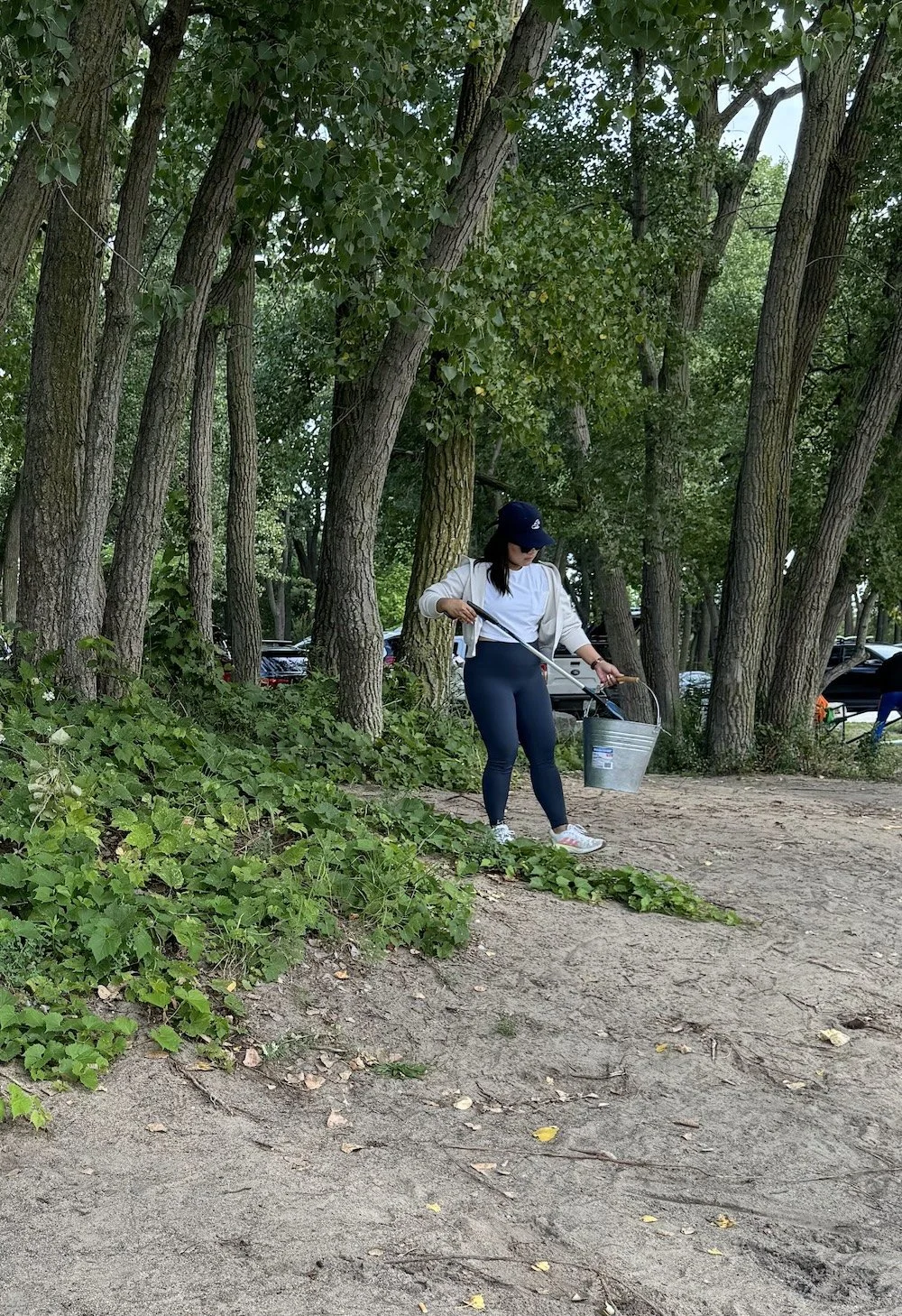 A woman wearing sunglasses, a baseball cap, a white jacket, and dark leggings collecting trash with a grabber and a metal bucket in a wooded park.