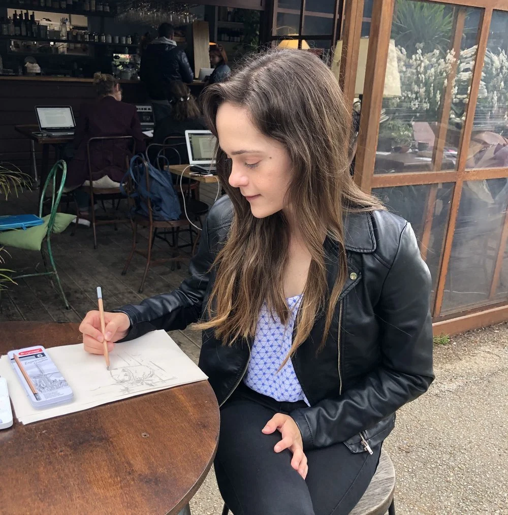 A young woman with long brown hair, wearing a black leather jacket, is sitting at a wooden table in a cafe, sketching in a notebook with a pencil. The cafe has large windows and several people in the background using laptops and dining.