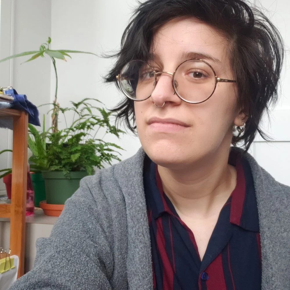 A person with black, messy hair, wearing glasses, a septum piercing, earrings, a gray cardigan, and a dark striped shirt, sitting in an indoor setting with plants and furniture in the background.