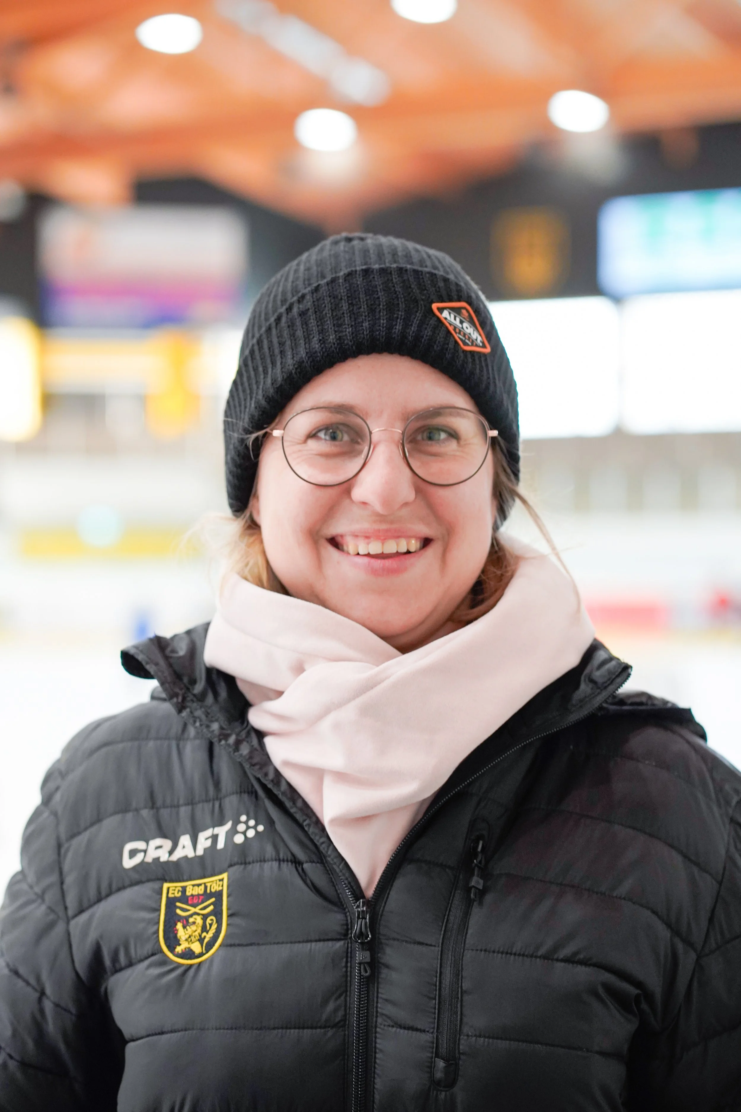 Ein junger Eishockeyspieler in grauem Trikot und eine Frau in einer schwarzen Jacke auf einer Eisbahn, beide lächeln in die Kamera.