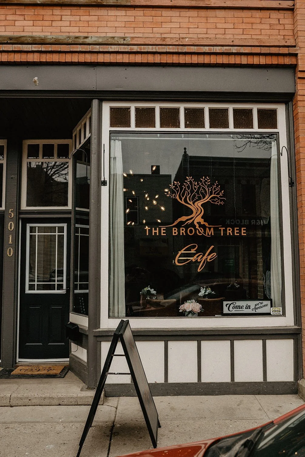 The storefront of The Broom Tree Cafe, with a large window displaying the cafe's logo of a tree, and a sign in the window that says "Come in, we're awesome."