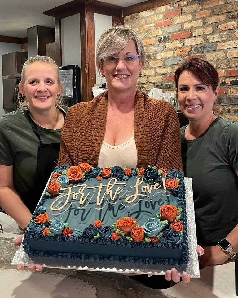 Three women smiling and holding a decorated cake with the words 'Jori the Love' on it, inside a room with a brick wall and kitchen background.