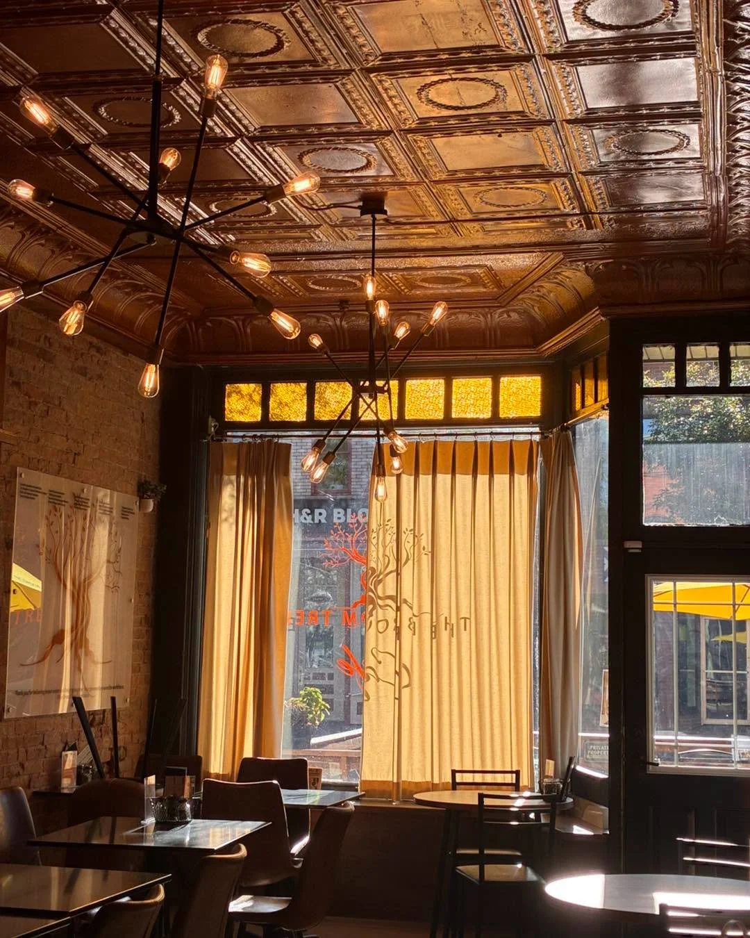 Interior of a cozy cafe with warm sunlight coming through large windows with yellow curtains, vintage patterned tin ceiling, modern black chandeliers with exposed bulbs, brick wall, and wooden tables and chairs.