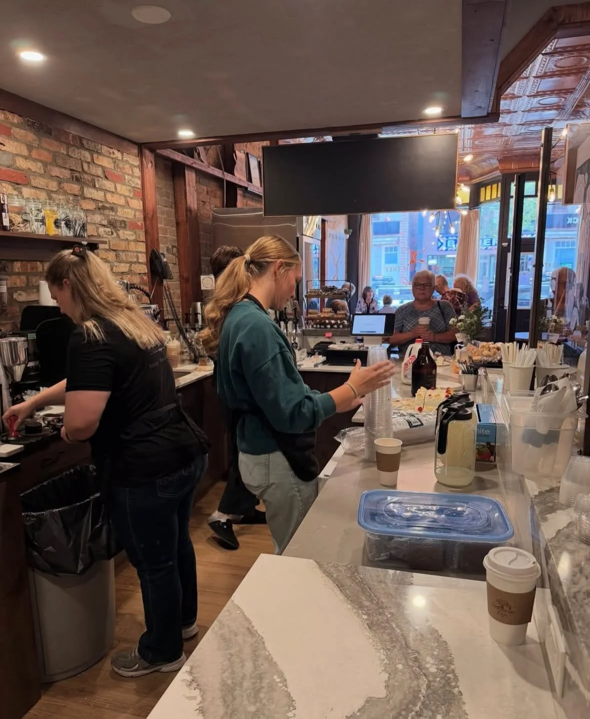 Interior of a coffee shop with two baristas and several customers, brick walls, and large windows.