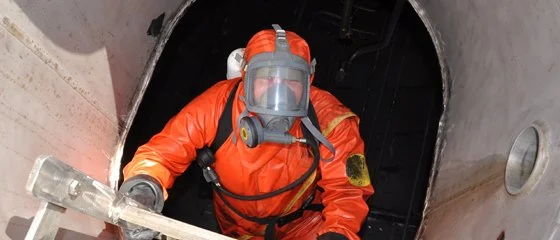 A worker in protective suit and a respirator climbing out of a large industrial tank.