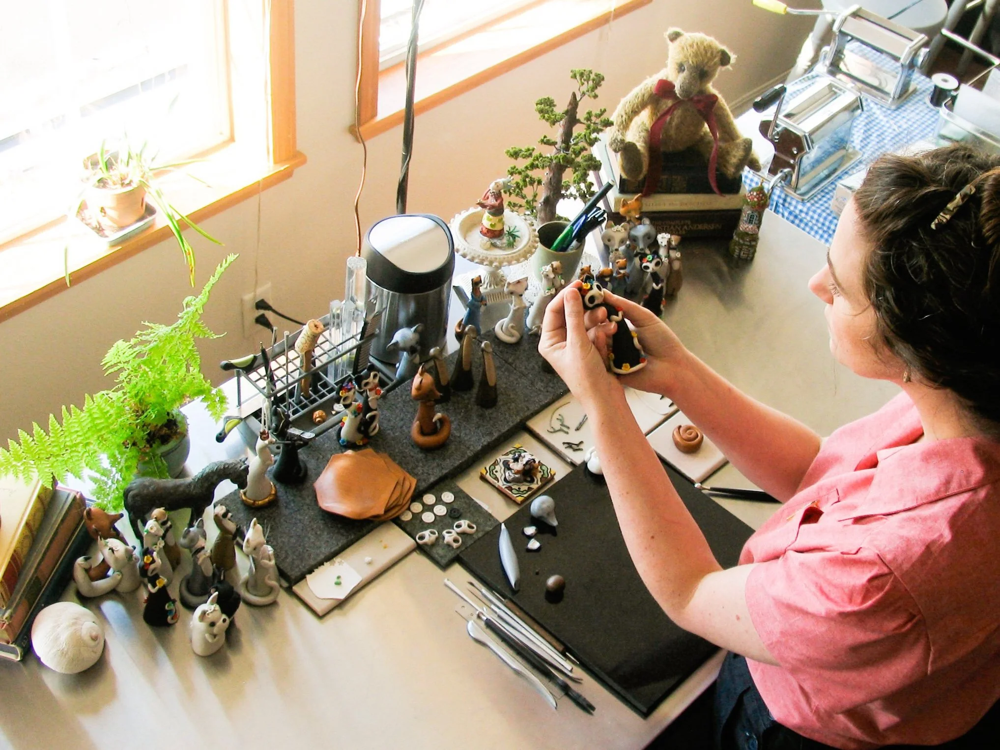 A woman at a worktable arranging handcrafted animal figurines, with various tools, plants, and decorative items on the table, creating detailed animal art.