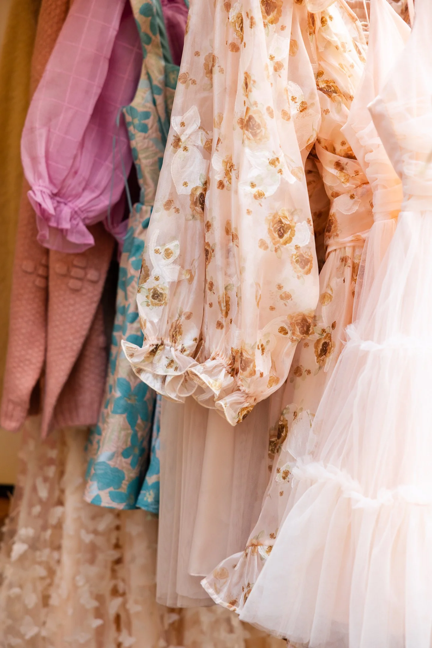 Close-up of hanging dresses, including floral and pastel-colored fabrics with ruffled sleeves and tulle skirts.