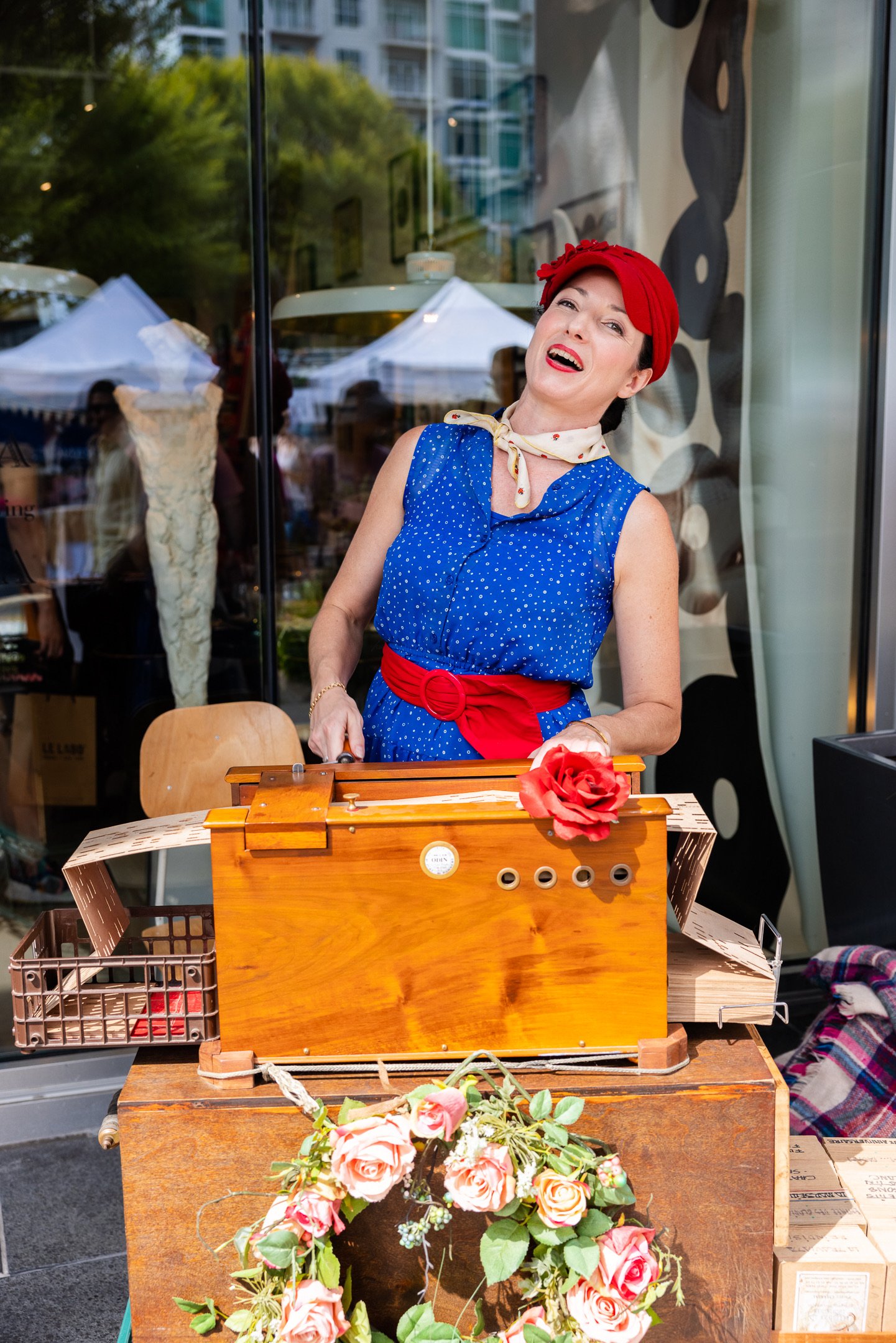 A woman dressed in 1950s style with a blue polka-dot dress, red headscarf, and red belt, standing behind a vintage wooden toy piano decorated with a red flower, outside a storefront with reflections of trees and tents in the window.