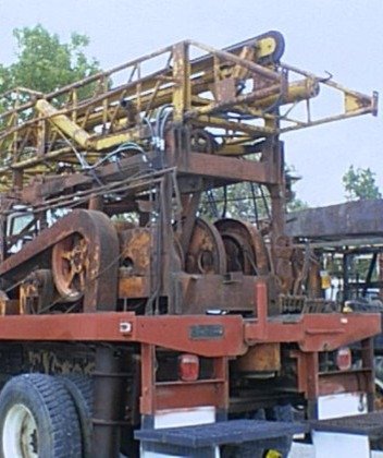Rusty industrial or construction machinery mounted on a trailer, with large gears and a yellow boom arm, set outdoors with trees in the background.