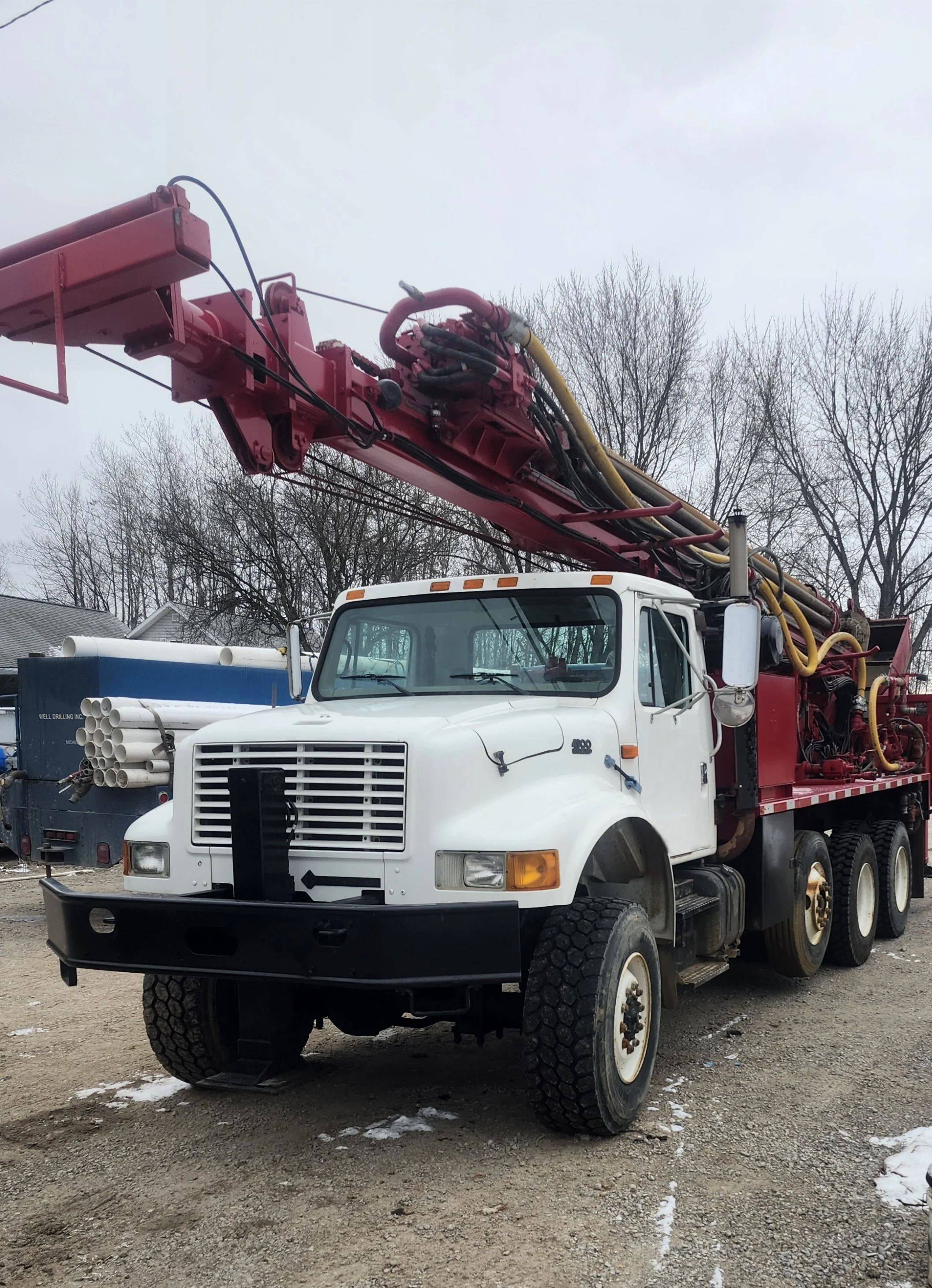 A large white and red industrial drilling truck with a tall pink drill mounted on the back. The scene is outdoors on a gravel surface with leafless trees and cloudy sky in the background.
