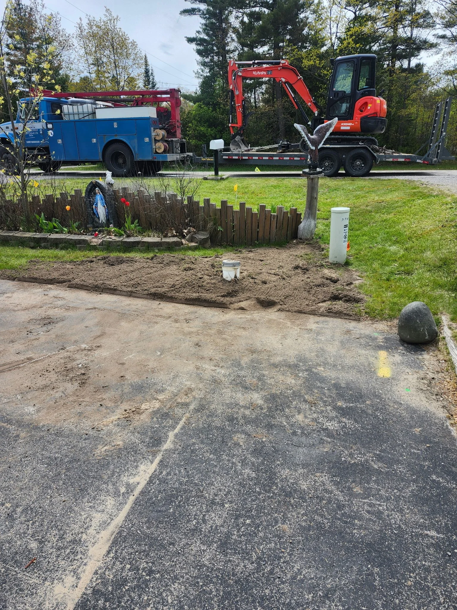 A small garden with a religious statue and flowers, a white pipe sticking out of the ground, and a decorative eagle sculpture. In the background, a red and black mini excavator on a trailer and a blue utility truck parked on the street, behind a tree