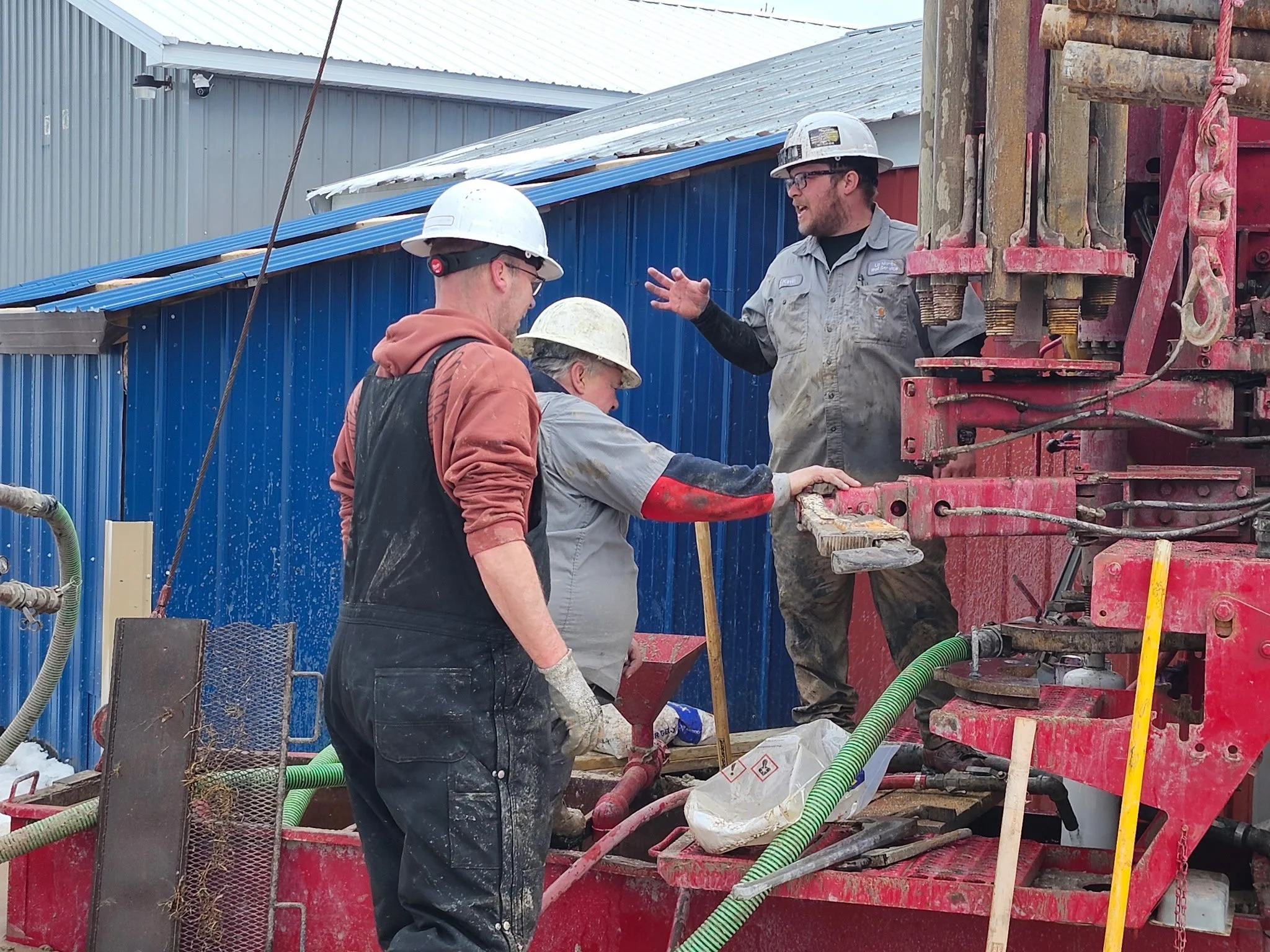One driller and 2 helpers  wearing hard hats and work clothes discussing next steps on a drilling site with machinery and well materials.