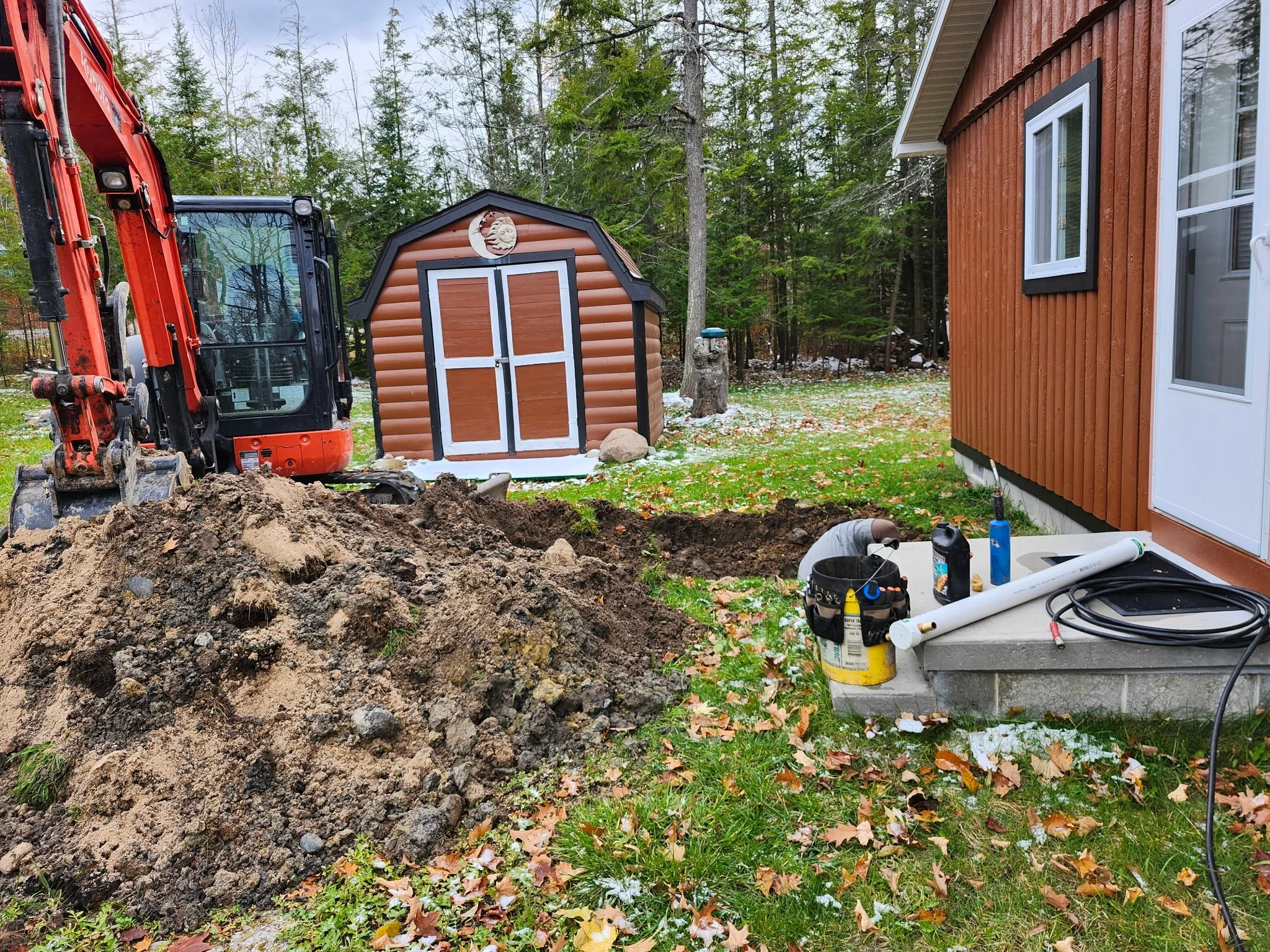 Backyard well hook up site with a small excavator digging near the house, a pile of dirt, a person working in a hole, a shed in the background, a wooded area with trees, and autumn leaves on the ground.