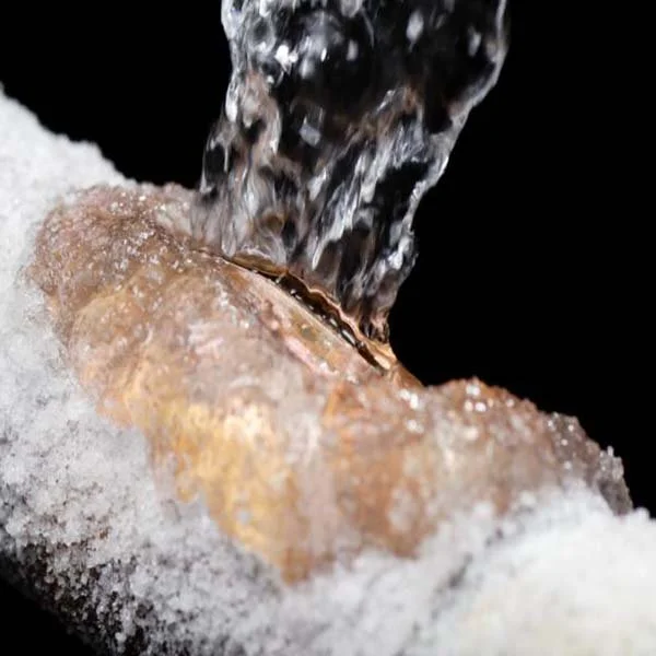 Close-up of a froze and burst copper pipe, with water pouring over it and surrounded by ice.