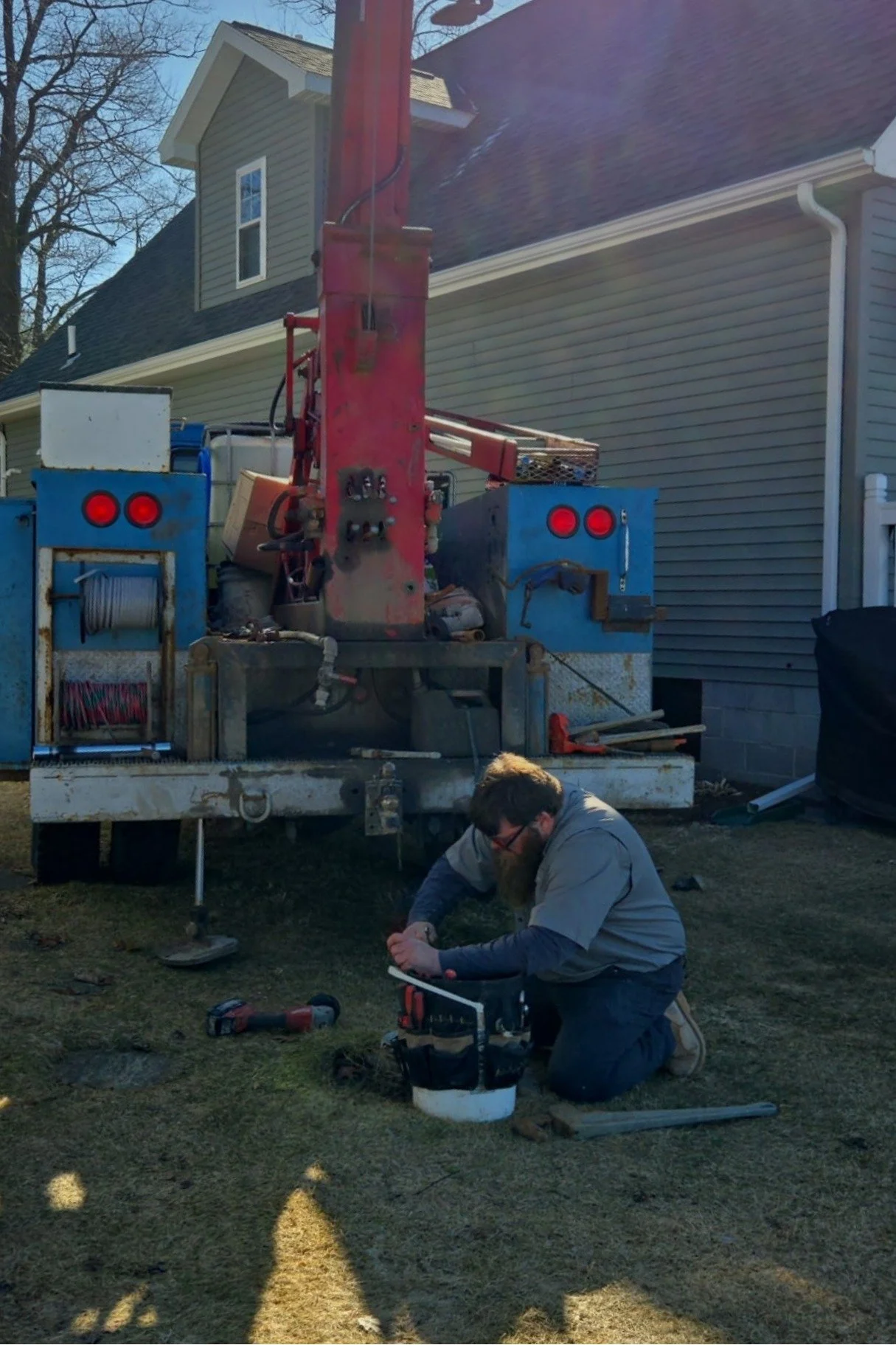 A man working on a water well outdoors, kneeling on the grass in front of a service truck with equipment and supplies.
