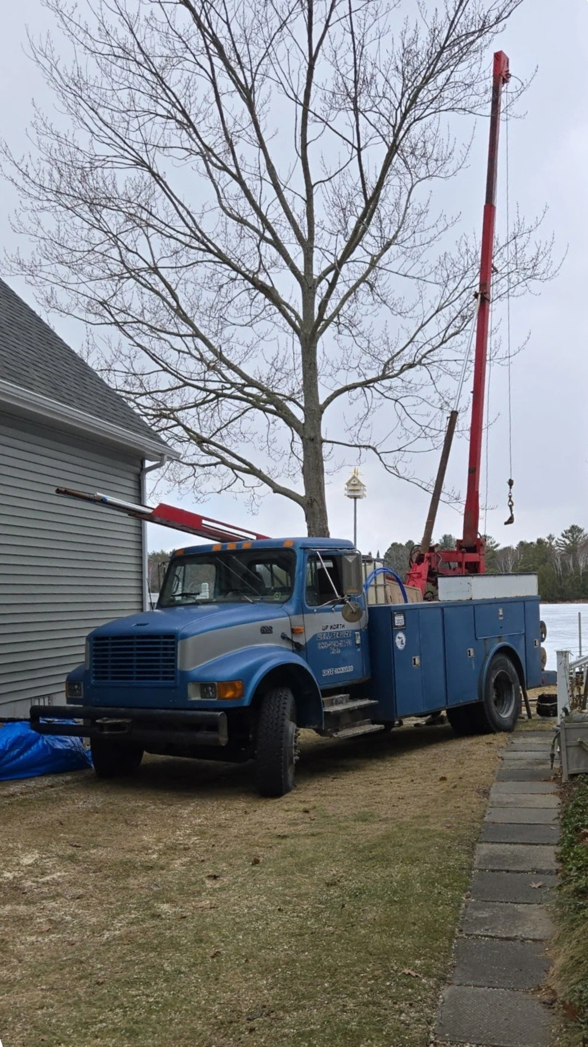 A blue service truck with a pump hoist attachment parked on a lawn next to a house, with a large leafless tree in the background.