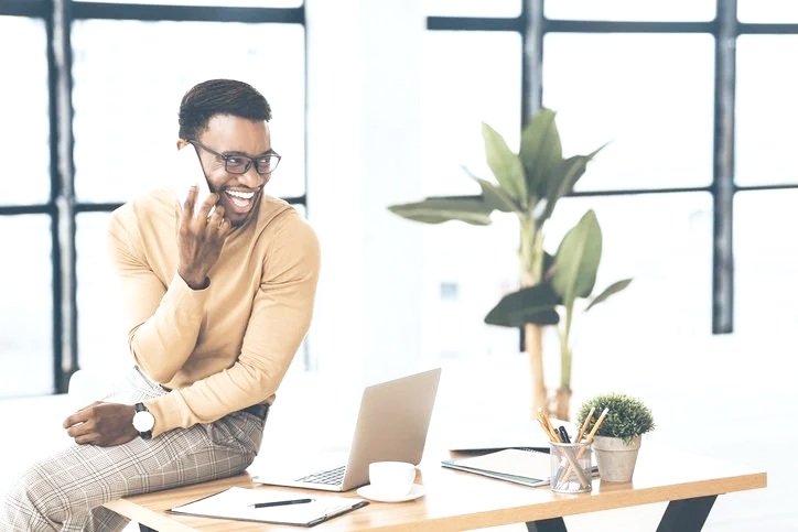 A smiling man with glasses sitting on a desk in a bright office, talking on a cellphone, with a laptop, coffee cup, notebooks, and office supplies on the desk, and a large green plant in the background.