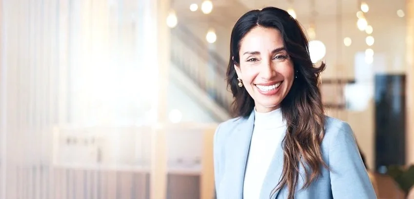 Portrait of a smiling woman with long dark hair, wearing a light blue blazer and white top, in a modern indoor setting.