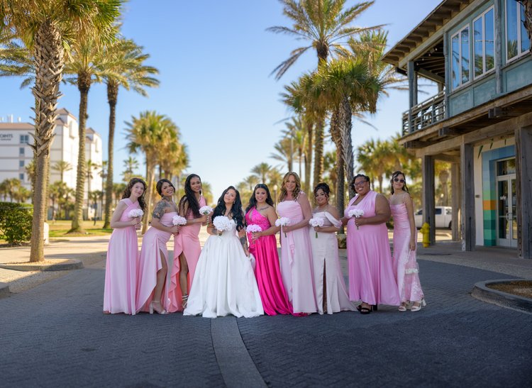 A bride and her bridesmaids in shades of pink pose along a palm-lined street on a clear day.