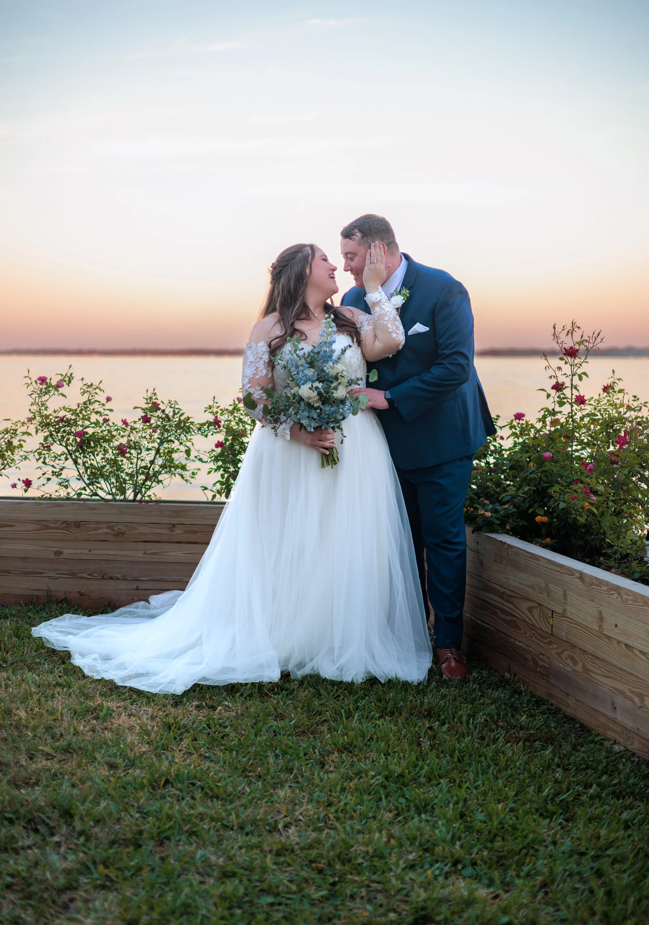 A bride and groom standing close together by a wooden planter filled with pink and red flowers, smiling at each other during sunset by a body of water.