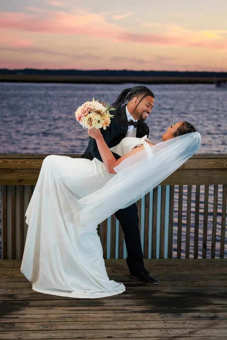 A bride and groom are dancing on a wooden pier by the water at sunset, with the groom lifting the bride while holding a bouquet of flowers.