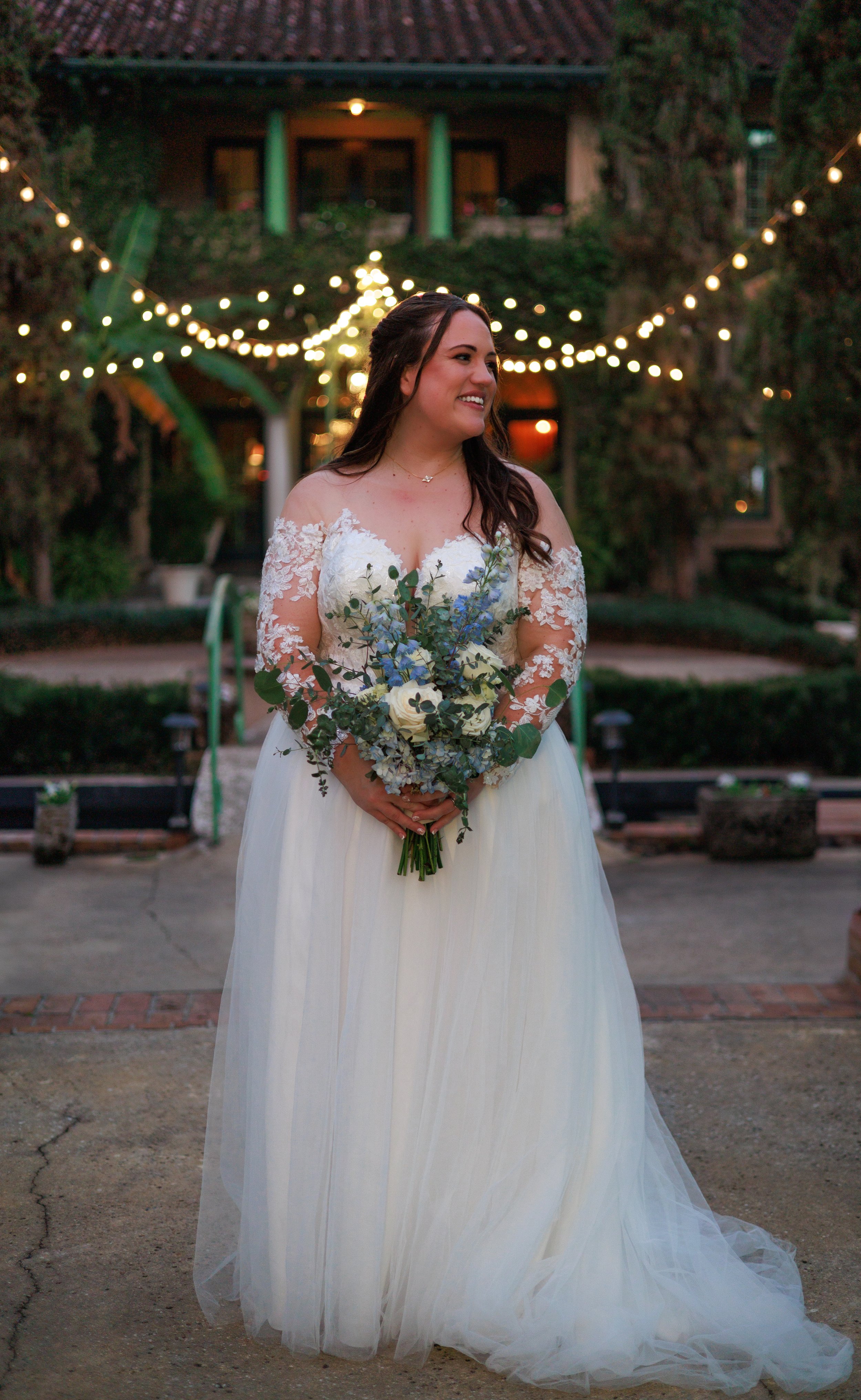 A bride in a white wedding dress holding a bouquet of flowers, standing outdoors at dusk with string lights overhead.