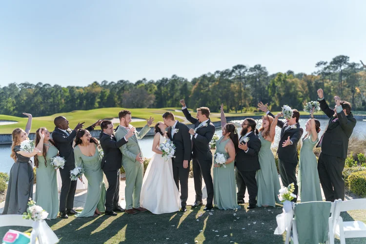 The wedding party applauds and cheers as the bride and groom embrace on a golf course lawn, bridesmaids in sage green and groomsmen in dark suits.
