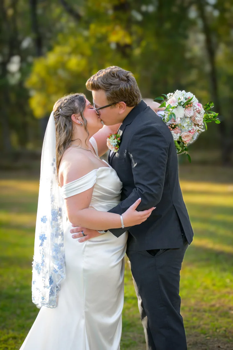 A bride and groom kiss in an open field at golden hour, her bouquet of soft pink and white blooms raised beside them.