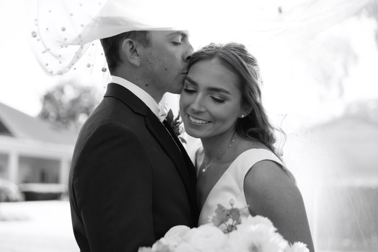 A black and white photo of a bride and groom standing closely, with the groom kissing the bride's forehead. The bride is smiling with her eyes closed, wearing a wedding dress and earrings, and holding a bouquet. The groom is dressed in a tuxedo with