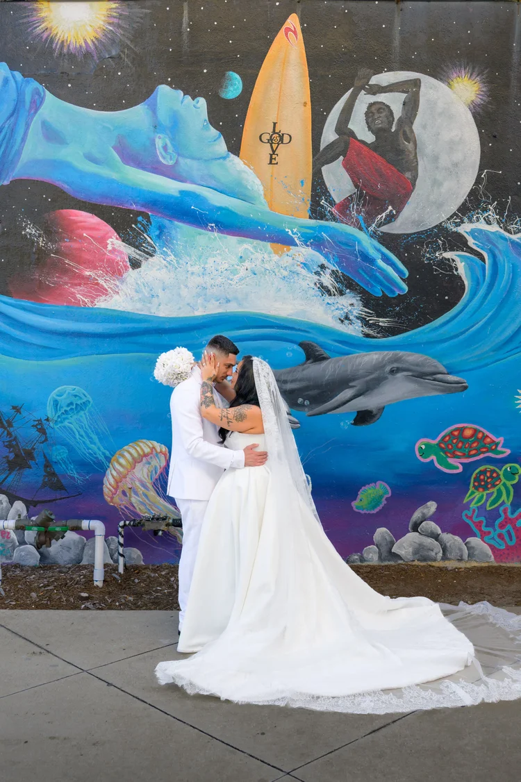 A bride and groom kiss in front of a large ocean-themed mural, her long train spread out across the pavement.