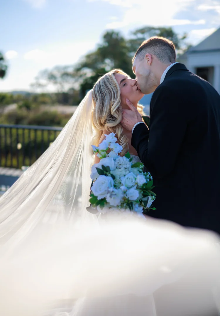 Bride and groom sharing a kiss outdoors, bride holding a bouquet of white roses, groom in a black suit, sunlight and trees in background.