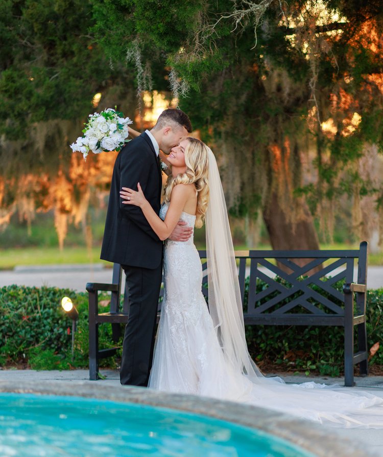 A bride and groom kiss beside a fountain beneath draping live oaks and Spanish moss, her long veil falling down the back of her gown.