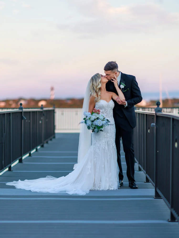 A bride and groom sharing a kiss on a rooftop during sunset, with the bride holding a bouquet of white and blue flowers, dressed in a lace wedding gown, and the groom in a black tuxedo.