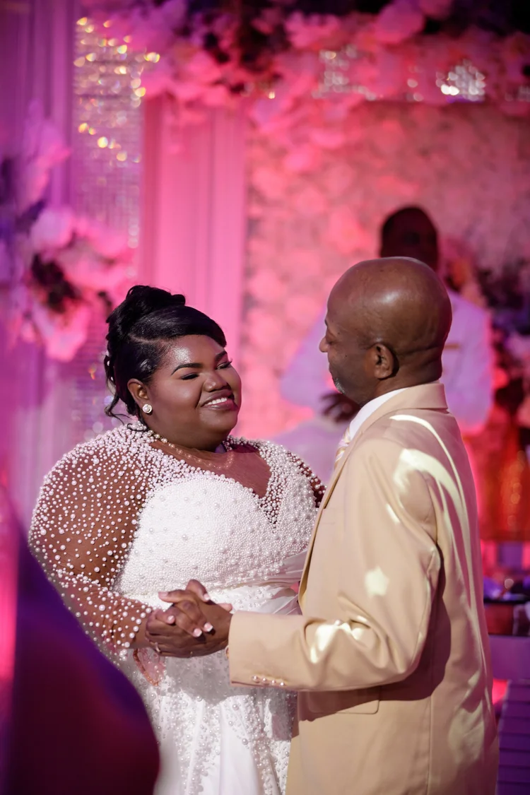 A bride laughs while holding her husband's hands during the reception, the room behind them washed in deep pink light.