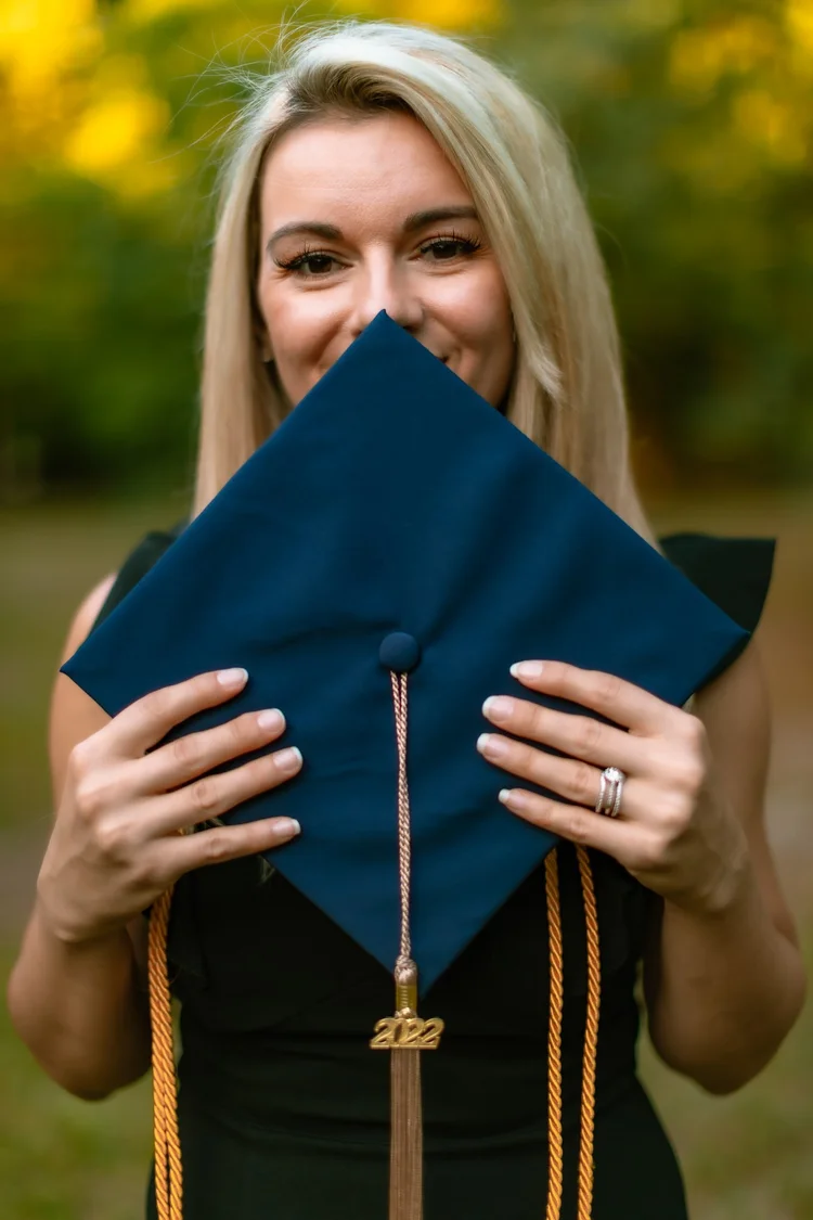 A graduate holds her navy cap and tassel in front of her face, smiling over the top of it.