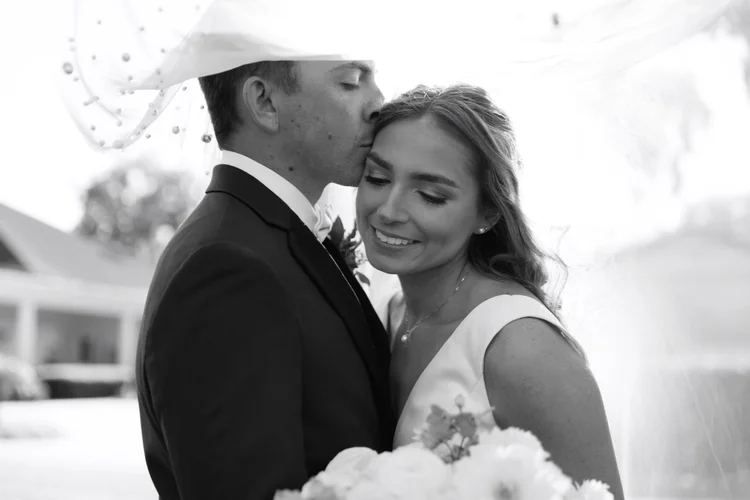 A black and white photo of a bride and groom standing closely, with the groom kissing the bride's forehead. The bride is smiling with her eyes closed, wearing a wedding dress and earrings, and holding a bouquet. The groom is dressed in a tuxedo with a bow tie and wearing an umbrella on his head, likely for protection from the rain.