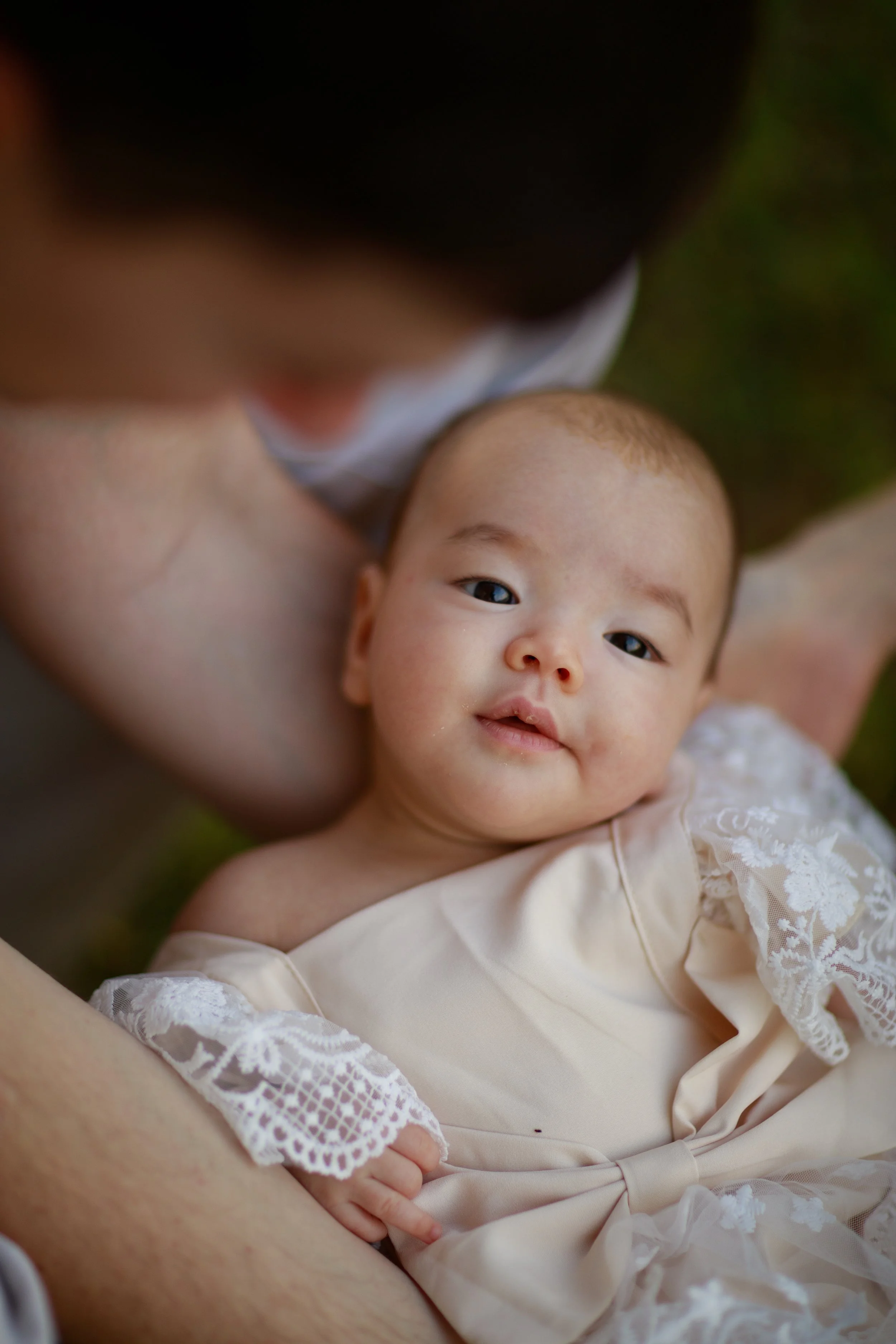 Close-up of a baby lying on an adult's arm outdoors, wearing a cream-colored dress with lace sleeves, looking at the camera.