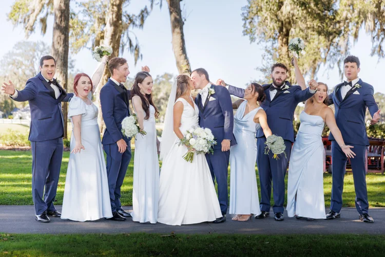 A group of people at a wedding, including a bride and groom kissing, surrounded by friends. The friends include men in suits and women in white dresses, some holding bouquets, all celebrating outdoors with trees in the background.