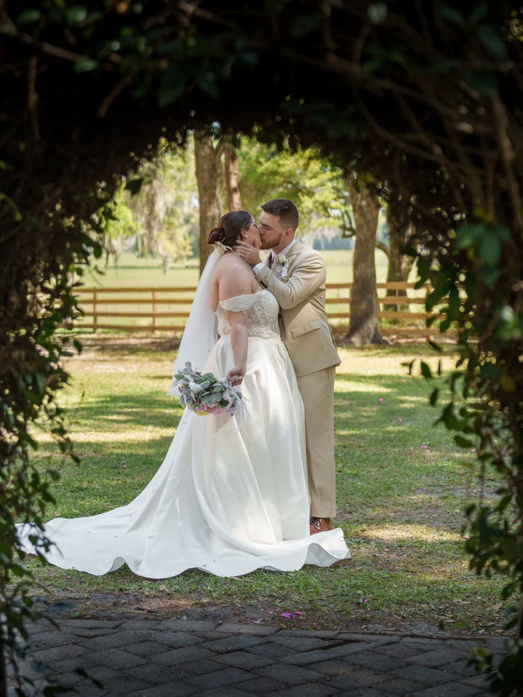 A bride and groom stand together inside a leafy garden archway, her gown pooling on the brick path behind her.