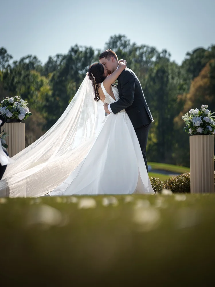 A bride and groom kiss at the end of the aisle between two tall floral arrangements, her long veil sweeping across the grass.