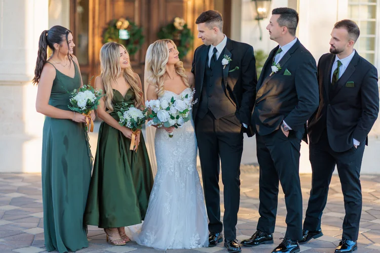 A wedding party lined up outside a stone-columned venue, bridesmaids in deep green gowns holding white bouquets beside groomsmen in black suits, with the bride and groom at the centre.